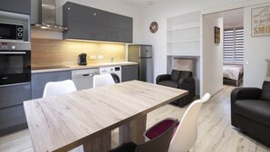 Cozinha moderna com eletrodomésticos embutidos, área de jantar com mesa de madeira e sala de estar com sofá e cama.