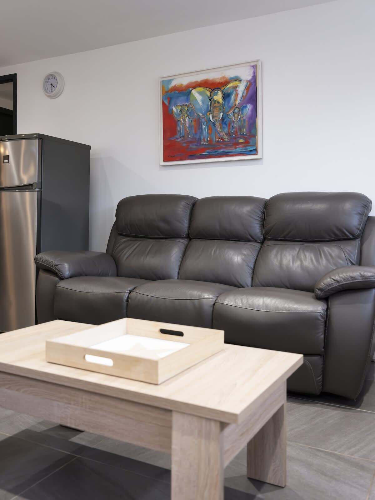 A living room with a black leather sofa, a wooden coffee table, a stainless steel refrigerator, and a colorful abstract painting on the wall.