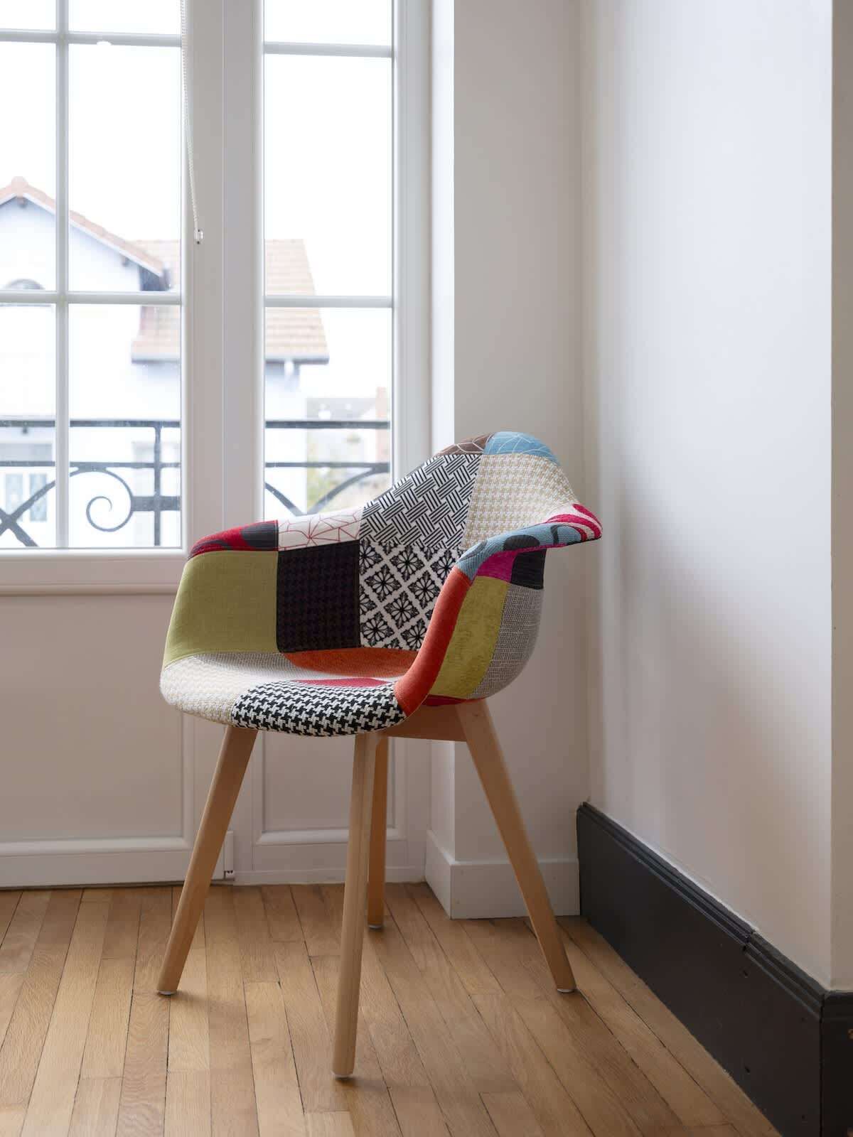 Une chaise au motif patchwork, avec des pieds en bois, et une fenêtre donnant sur des immeubles.