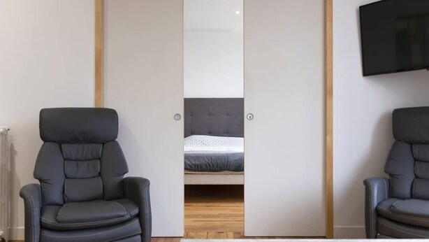 Une pièce comprenant un lit, deux chaises, un téléviseur et une porte de placard coulissante.