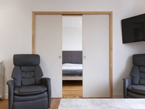 Ein Zimmer mit einem Bett, zwei Stühlen, einem Fernseher und einer Schiebetür zum Kleiderschrank.