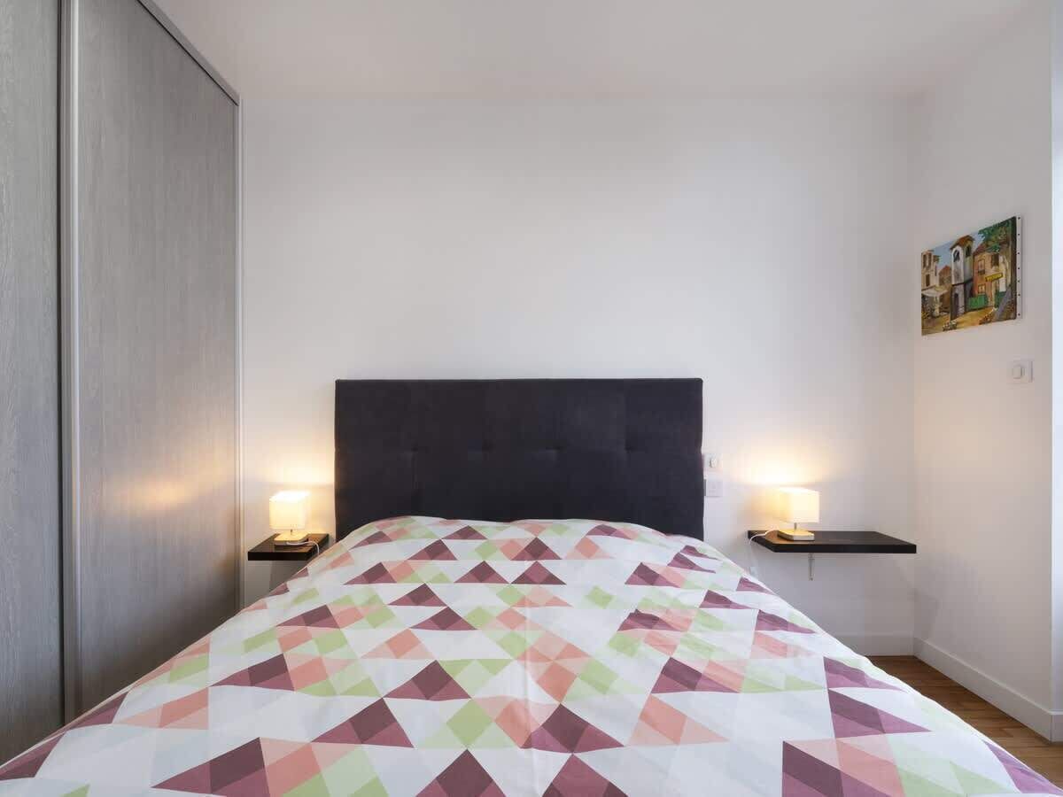Un lit avec une couvre-lit à motifs géométriques, deux tables de chevet avec des lampes, une tête de lit sombre, un tableau accroché au mur et une armoire.