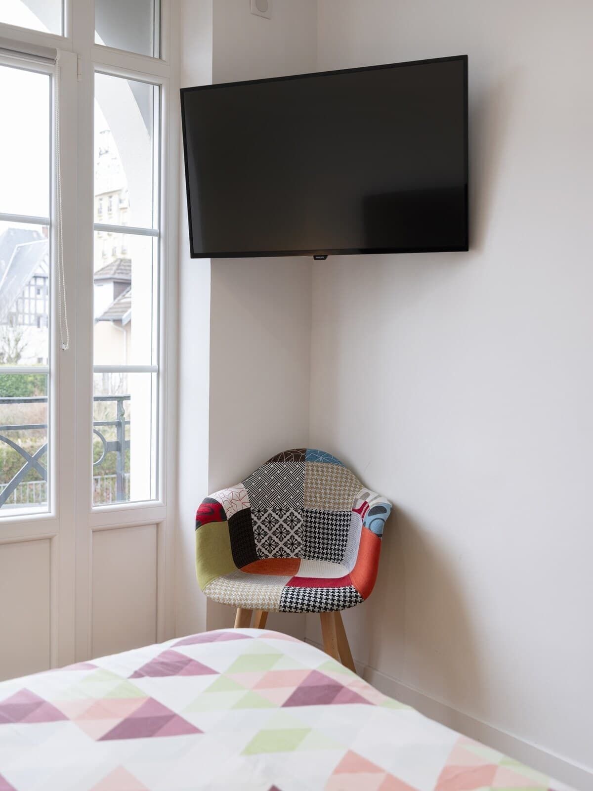 A room with a bed, a colorful chair, a TV mounted on the wall, and a window with a view of buildings.