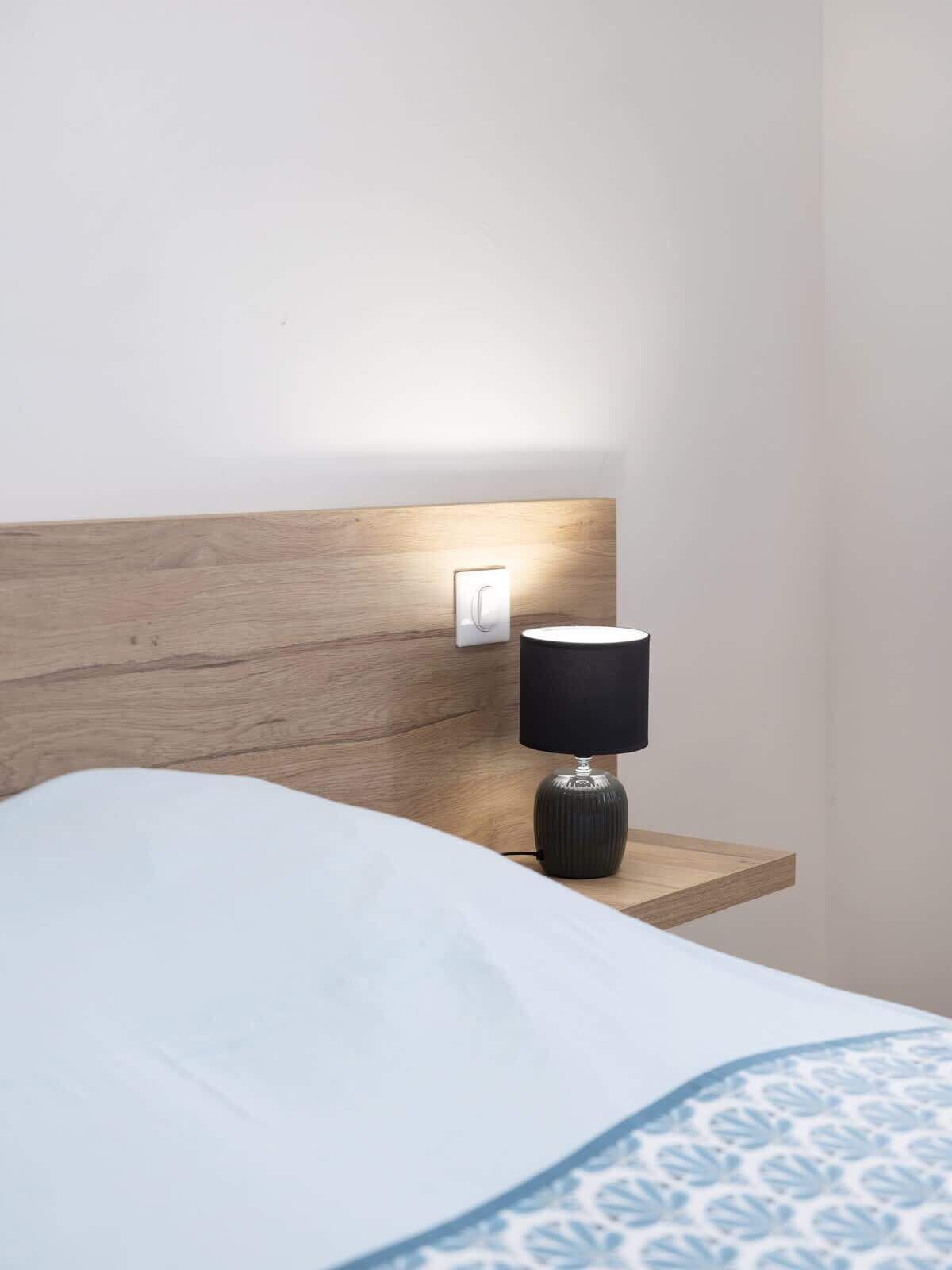 A bed with a white and blue patterned bedspread, a wooden headboard, a bedside table with a black lamp, and a wall-mounted light switch.