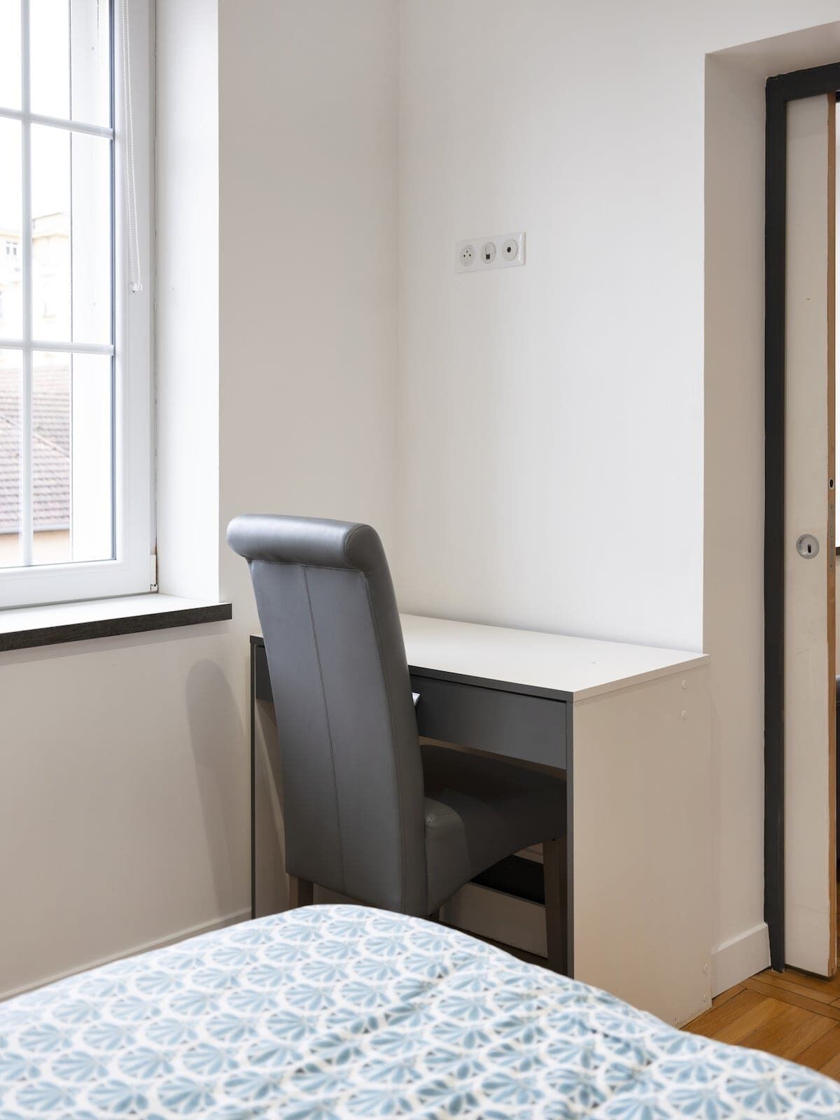 A bedroom with a bed, a desk, and a chair. There is a window and a door.