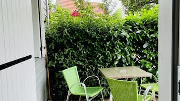 A wooden deck with two green chairs and a table, a white door, and a hedge in the background.