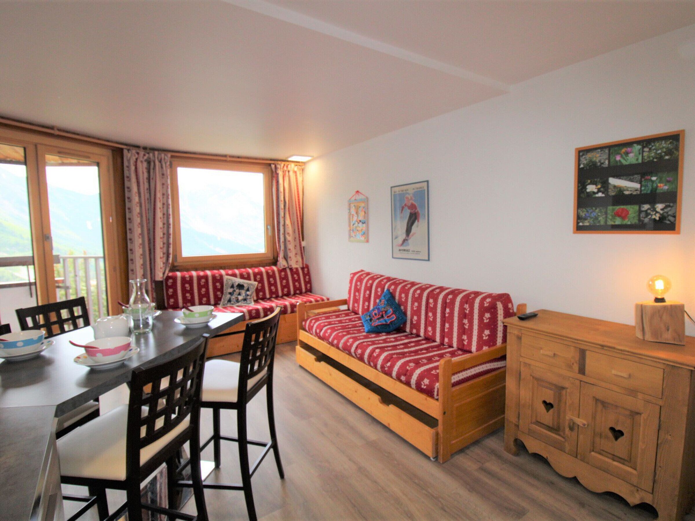 A cozy room with a wooden dining table, chairs, a sofa with patterned cushions, a cabinet with heart-shaped handles, and a window with curtains.