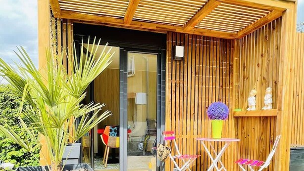 A wooden deck with a small table and chairs, a potted plant, and a building entrance with a glass door. - Cabane de Rafaël (Vittel)