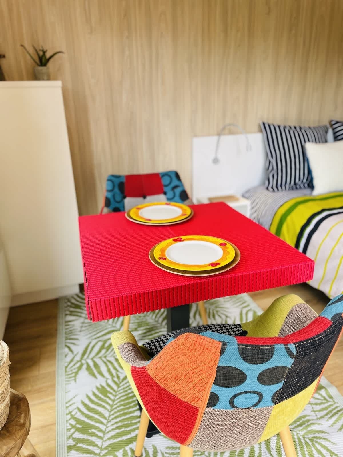 Une petite pièce aux couleurs vives, avec une table rouge, deux chaises et un lit agrémenté d’oreillers à rayures.