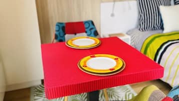 Une petite pièce aux couleurs vives, avec une table rouge, deux chaises et un lit agrémenté d’oreillers à rayures.