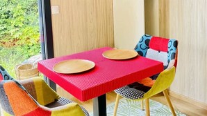 A modern dining area with a red table, colorful chairs, a wooden wall, and a view of greenery outside. - Cabane de Rafaël (Vittel)