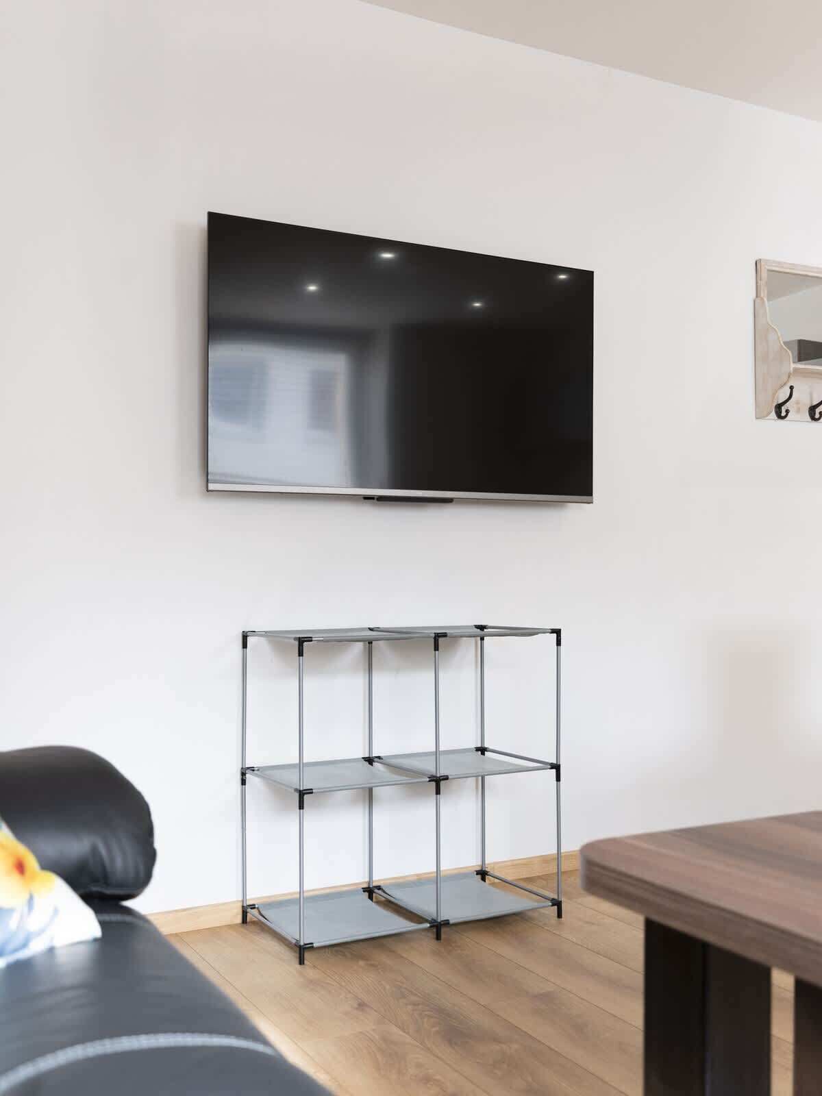 A modern living room with a mounted TV, a metal shelving unit, a wooden dining table, and a black leather sofa.