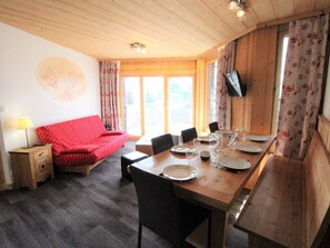 A cozy room with a red sofa, wooden dining table set with plates and glasses, and a TV mounted on the wall.