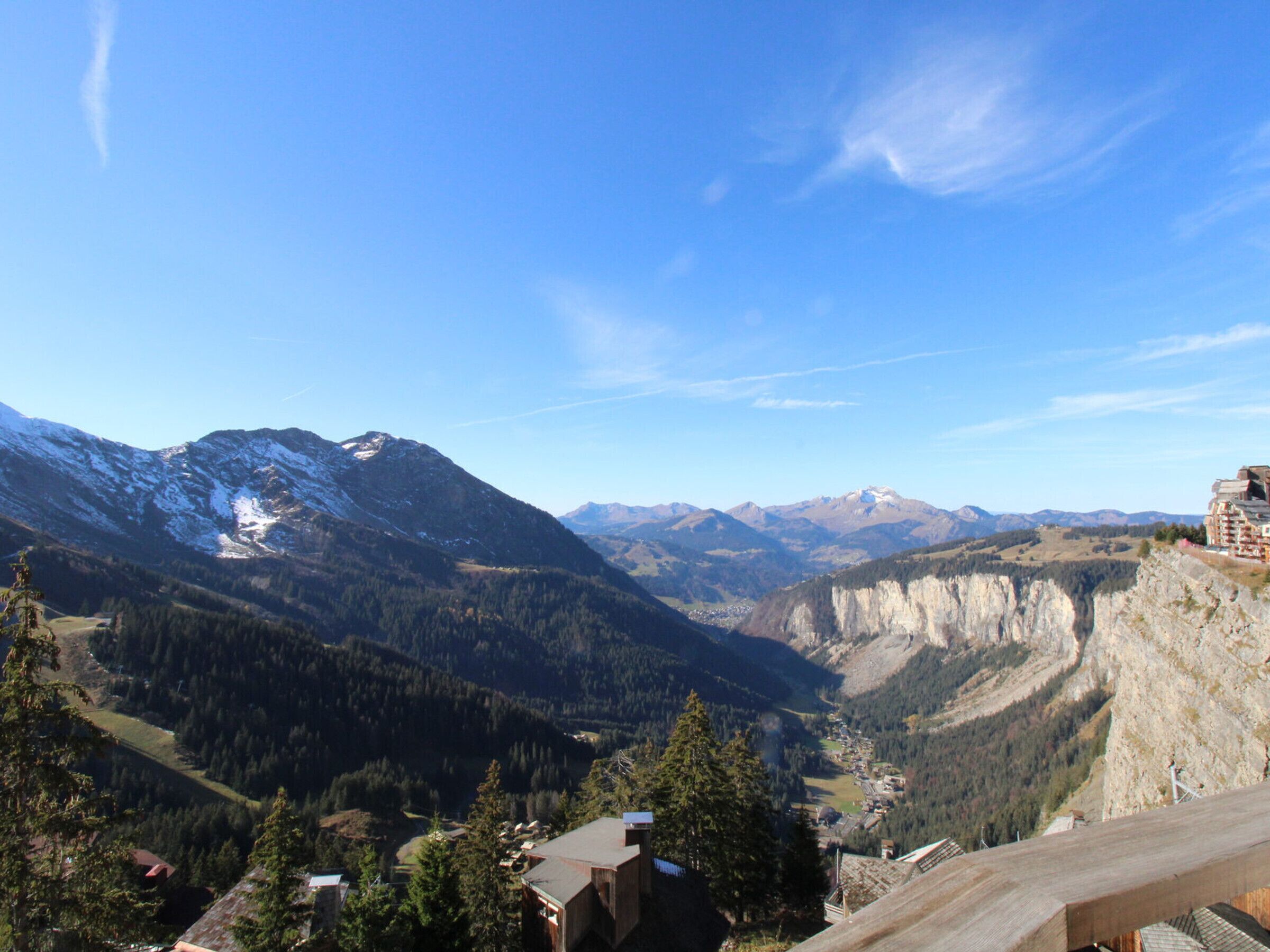 A mountainous landscape with a clear sky. There are buildings with flat roofs and a wooden structure in the foreground.