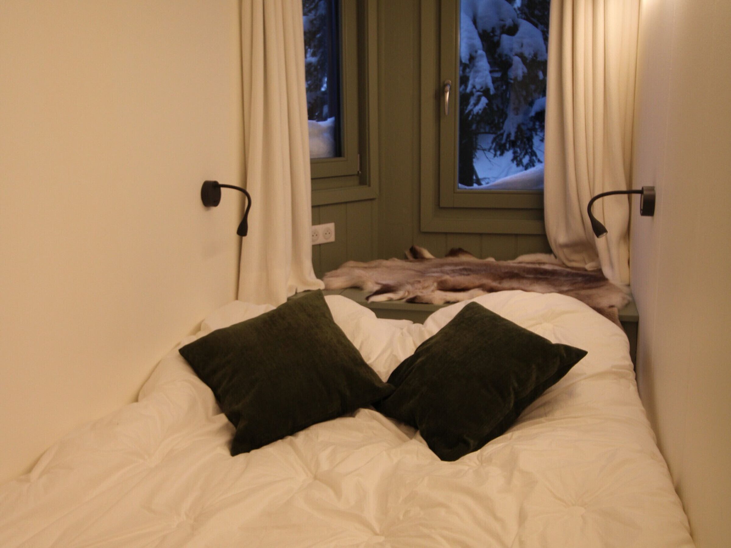 A bed with white bedding and two dark pillows, a window with curtains, and a view of snow-covered trees outside.