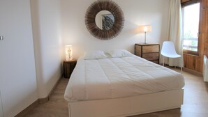 Une chambre avec un grand lit, une table de chevet avec une lampe, une chaise, un miroir rond et une fenêtre avec des rideaux.