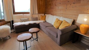 Un salon chaleureux avec un mur en bois, un canapé avec des coussins, un fauteuil moelleux blanc et deux petites tables en bois.