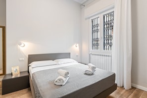 A neatly made bed with two pillows and two rolled towels, a bedside table with a picture frame, a window with decorative metal bars, and a radiator. - GuestHost - Comfortable 40 sqm apartment that can accommodate up to 4 people. Located on the ground floor (steps to access) of a building in Maragliano / Redi district, a lively neighborhood well connected to the historic center by public transportation, (Firenze)