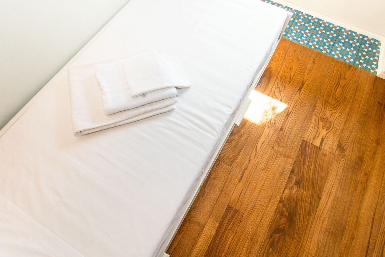 Una cama bien hecha con sábanas blancas y una toalla doblada encima, suelo de madera y un borde estampado al pie de la cama.