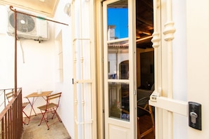 Exterior blanco con una mesa y sillas de madera, una puerta con ventanal y un equipo de aire acondicionado.