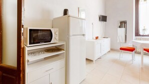 Une cuisine compacte équipée d’un four à micro-ondes, d’un réfrigérateur, de meubles blancs, d’une petite table et d’une fenêtre avec des rideaux.
