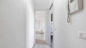 Un couloir étroit avec un mur blanc, un téléphone fixé sur le côté droit et une porte donnant sur une pièce où se trouve une chaise blanche.