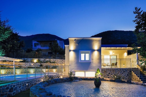 A modern house with a stone wall, a pool, and a well-lit patio area.