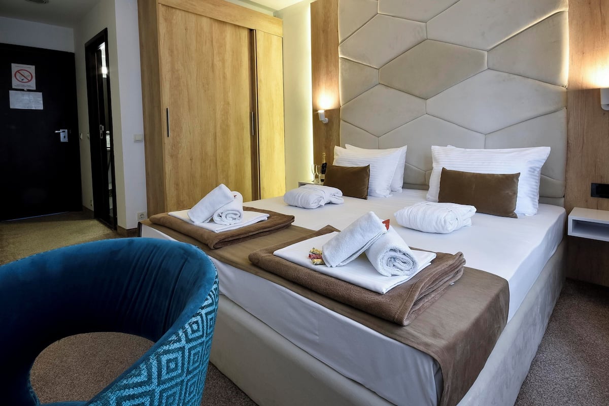 Quarto de hotel com duas camas, um guarda-roupa de madeira, uma cadeira azul e cabeceira com padrão geométrico.