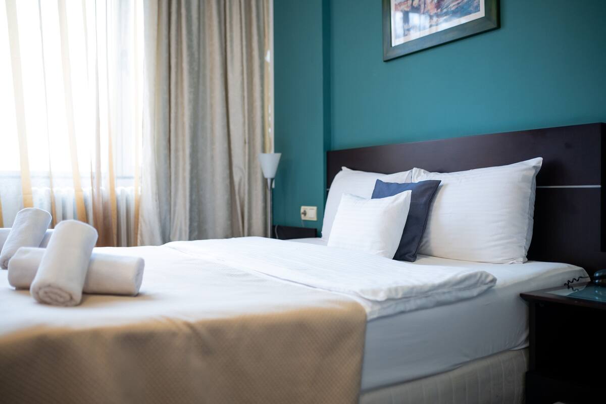 Uma cama bem arrumada com lençóis brancos e cabeceira escura, uma luminária em uma mesa de cabeceira e uma janela com cortinas.