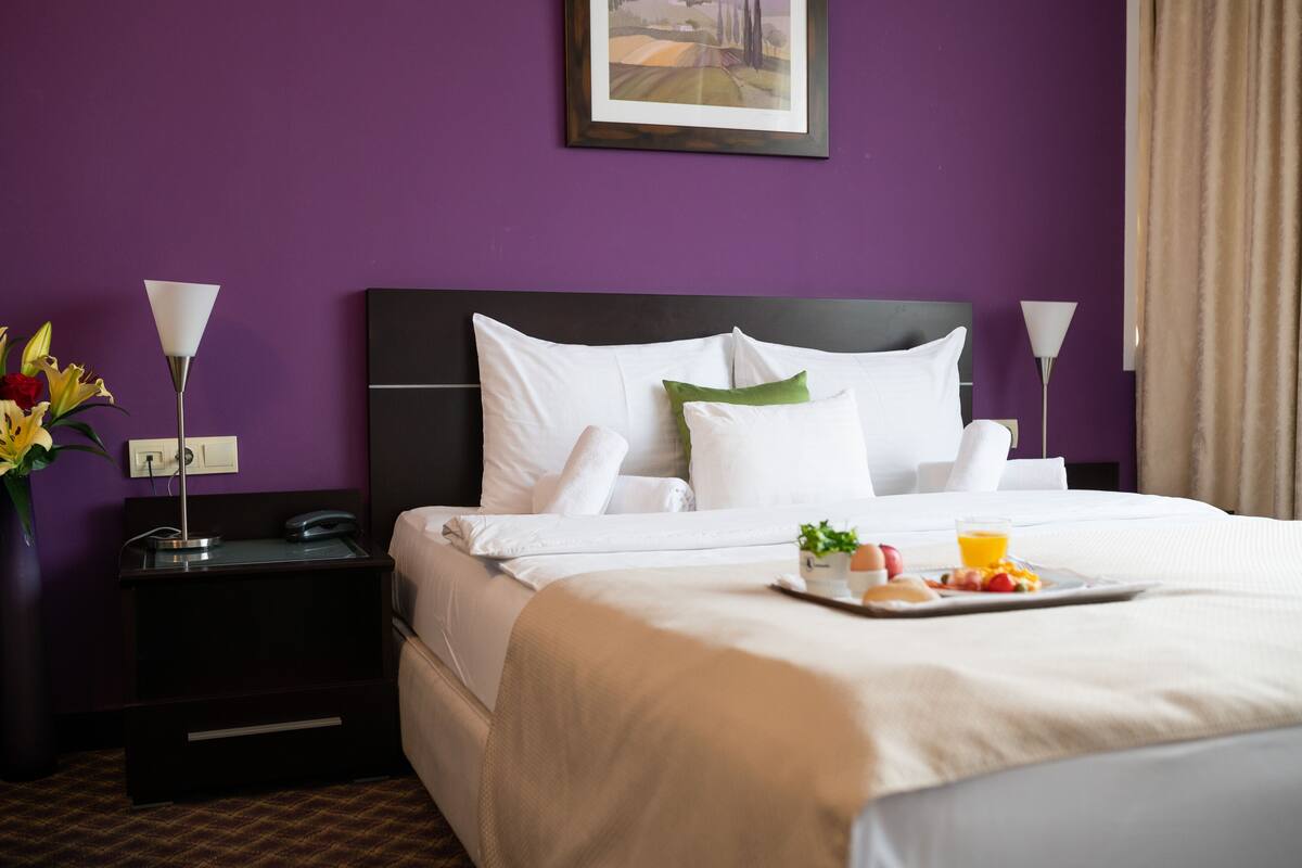 Quarto de hotel com cama, roupa de cama branca, bandeja com café da manhã, mesa de cabeceira com telefone e um vaso de flores.