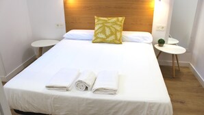 Een netjes opgemaakt bed met witte lakens en één decoratief kussen, samen met twee opgerolde handdoeken.