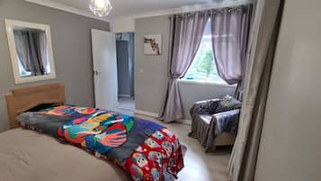 Un dormitorio con cama, ventana con cortinas, puerta y espejo fijado a la pared.