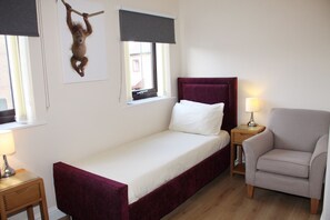 Una camera piccola con un letto singolo, un poggiatesta rosso, un comodino con una lampada e una sedia.