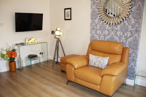 A living room with a leather armchair, a glass coffee table, a flat-screen TV, a floor lamp, and a decorative mirror.