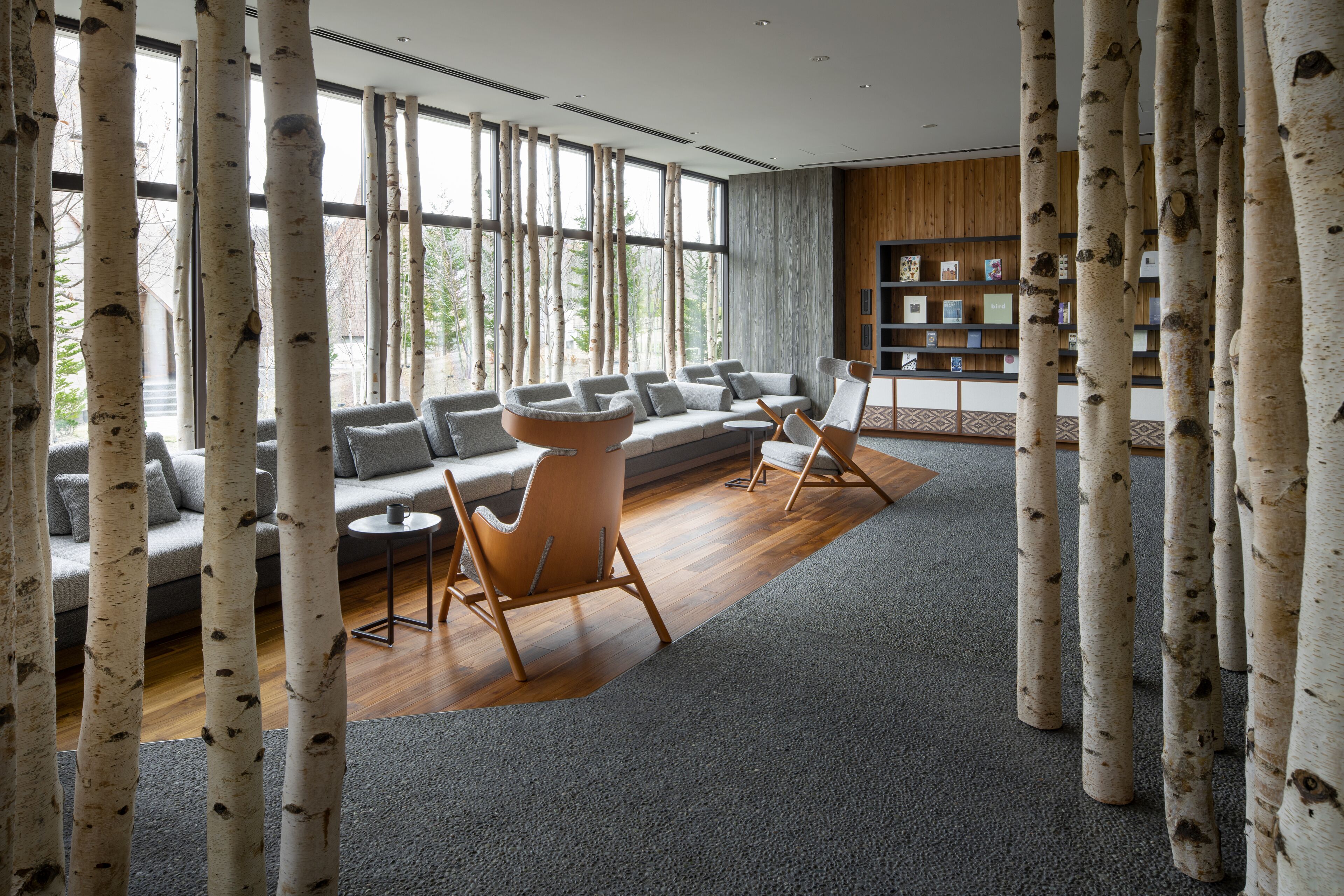 A modern living room with a large sofa, two armchairs, and a coffee table. There are birch tree-like columns in the foreground, and a bookshelf with books and decorative items in the background.
