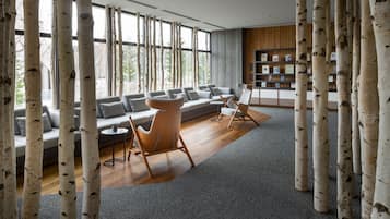 A modern living room with a large sofa, two armchairs, and a coffee table. There are birch tree-like columns in the foreground, and a bookshelf with books and decorative items in the background.