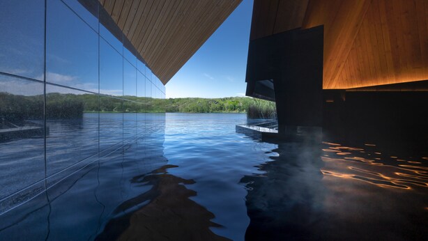 Ein modernes Gebäude mit Glasfassade, das eine Landschaft und einen Poolbereich mit Holzboden reflektiert.
