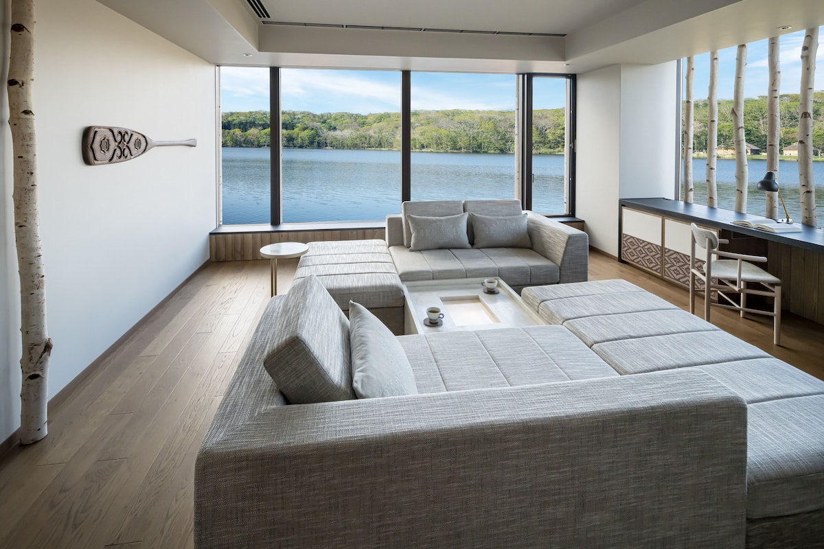 Ein modernes Wohnzimmer mit einem großen Fenster mit Blick auf einen See, einem grauen Ecksofa und einem kleinen Tisch mit einer Tasse darauf.