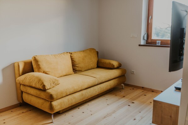 A room with a mustard-colored sofa, a wooden floor, a window with a view, and a flat-screen TV mounted on the wall.