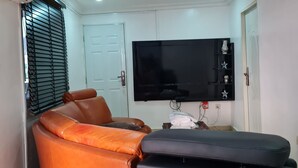A living room with a large flat-screen TV mounted on the wall, a brown leather sofa, and a black ottoman. - Beautiful Suits Apartment Near Airport Lagos (Lagos)