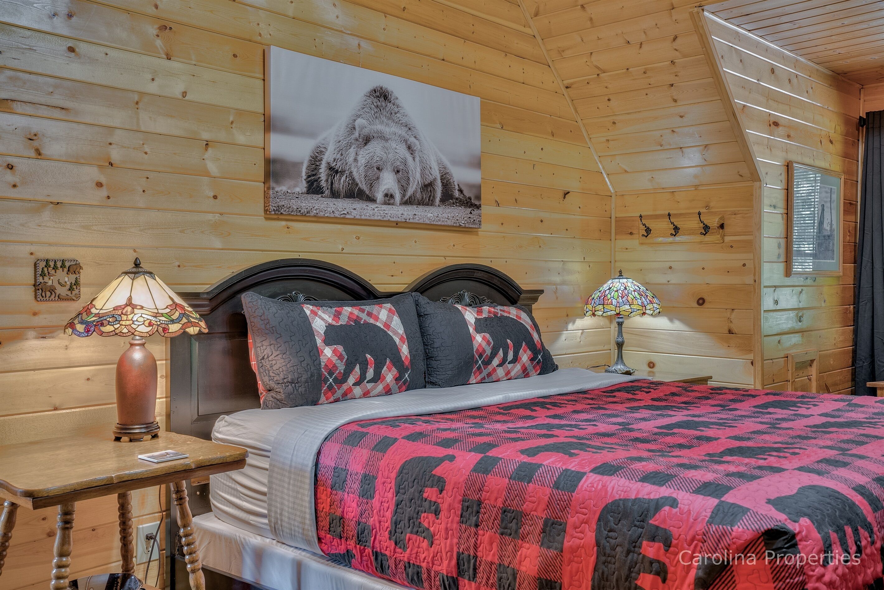 A cozy cabin-style bedroom with a wooden ceiling, a large bed with a red and black quilt, two bedside lamps, and a bear-themed pillow.