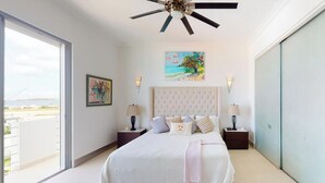 A bedroom with a large bed, a ceiling fan, a painting, and a view of the sea.