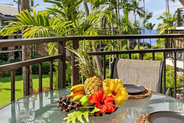 Un área de comedor al aire libre con ambiente tropical, que incluye una mesa de vidrio, individuales de mimbre y un arreglo de frutas con piña, plátanos, mango y flores de hibisco.
