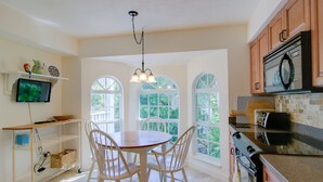 A kitchen with a dining table, chairs, a TV, and a view of greenery through large windows.