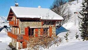 Ein Chalet mit geneigtem Dach und Holzbalkon, umgeben von verschneiter Landschaft und vereinzelten Bäumen.