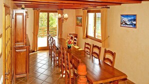 Ein Essbereich mit einem langen Holztisch, Stühlen und einem Blumenstrauß. Es gibt Fenster mit Vorhängen, eine Chandelier und ein Gemälde an der Wand.