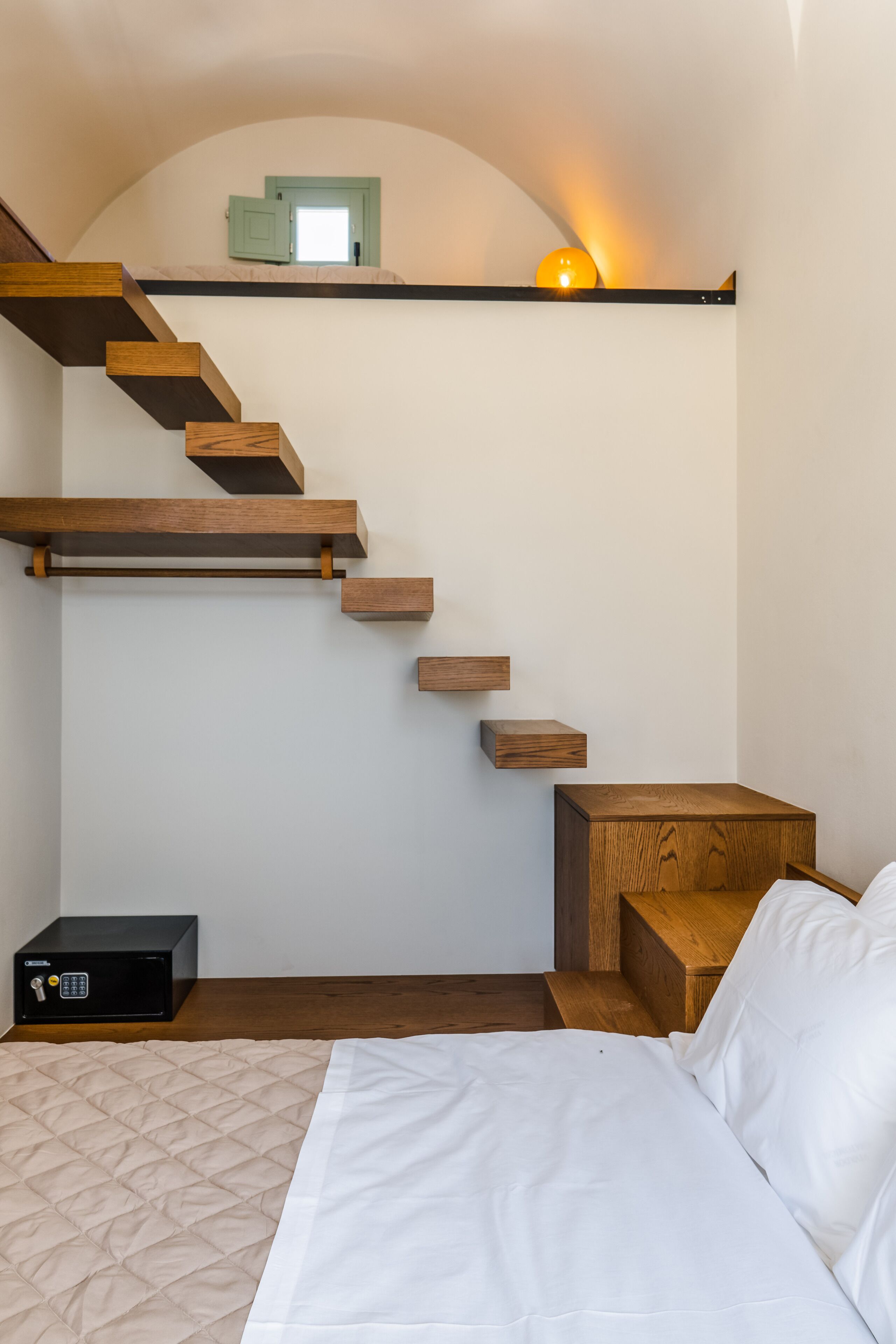 A bedroom with a bed, a bedside table, a nightstand, a wooden ladder staircase, and a safe.
