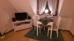 A small, cozy room with a television, a small table with chairs, a potted plant, and a window with curtains. - Cozy 2 room apartment (List)