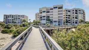 Une promenade en bois mène à un ensemble d’immeubles situés en bord de mer, dotés de nombreux balcons, sous un ciel dégagé.