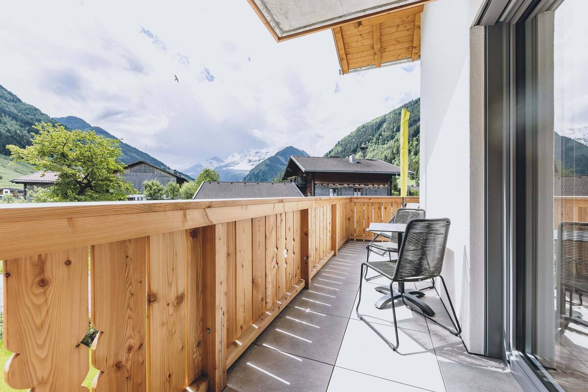 A wooden balcony with a view of mountains, a table, and chairs.